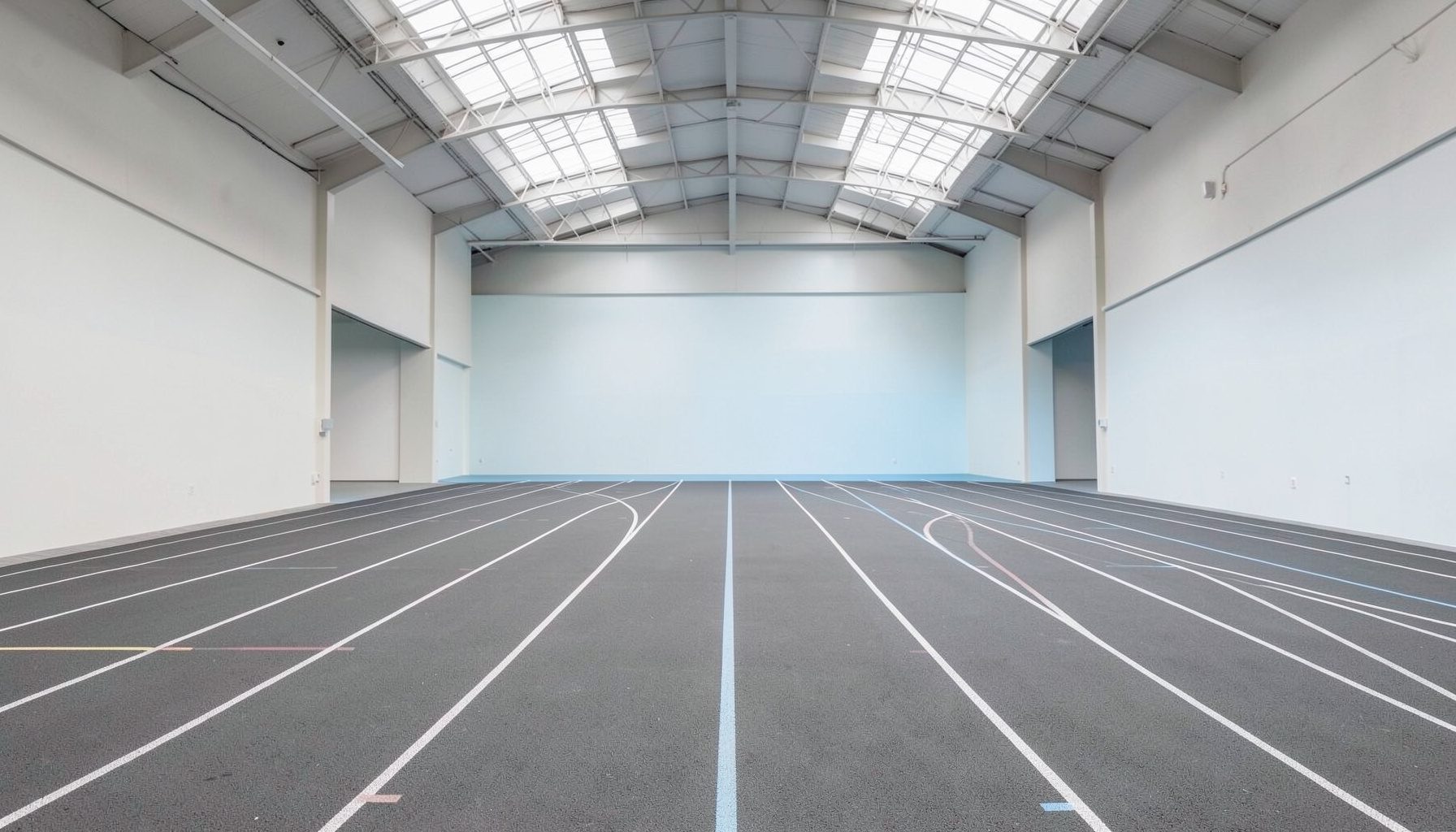 Clean architectural photograph with strong symmetry: a wide-angle view of an empty indoor running track with curved lanes and a matte rubber surface, lit by overhead skylights. The palette is muted gray, off-white, and pale blue, evoking discipline and quiet determination.