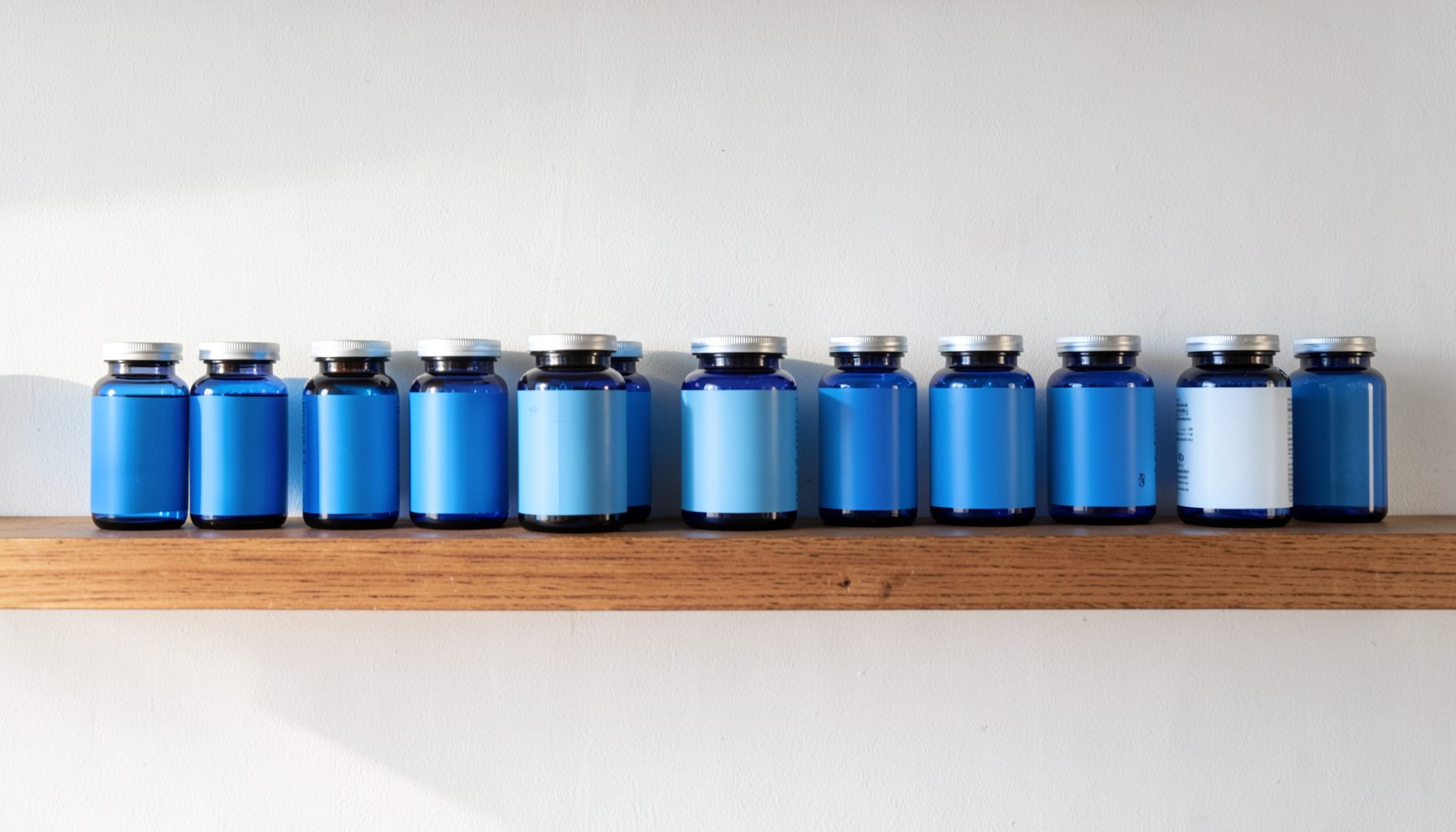 A neatly arranged lineup of supplement bottles on a wooden shelf against a white wall, with soft morning light casting gentle shadows.