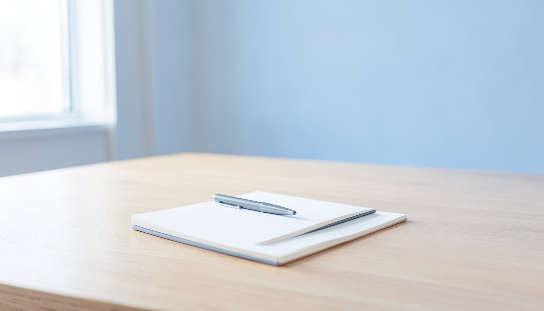 A clean, minimalist desk scene with a notepad and pen on a light wood surface, bathed in soft natural window light. The mood is calm, professional, and approachable.